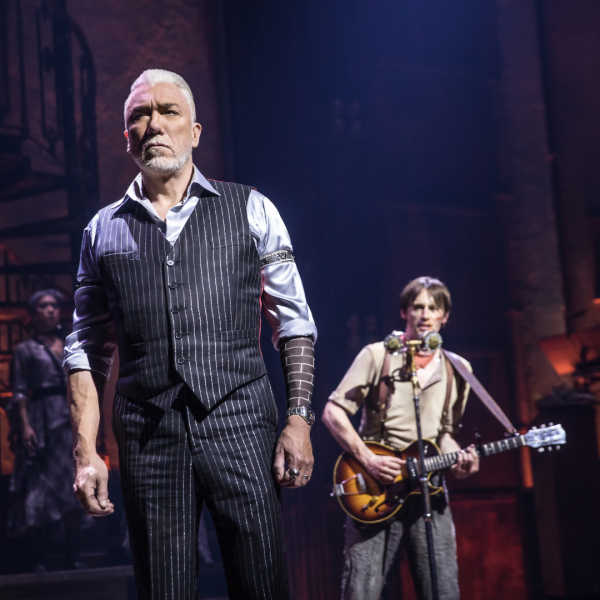 Photo of two main Hadestown leads - Hades at the forefront, and Orpheus playing a guitar at a mic stand
